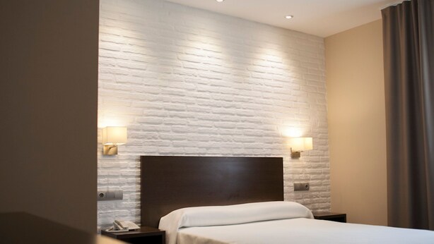 A hotel room with a white brick wall, a wooden headboard, a bed with white bedding, two bedside lamps, and a nightstand with a phone.