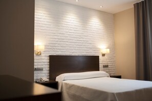A hotel room with a white brick wall, a wooden headboard, a bed with white bedding, two bedside lamps, and a nightstand with a phone.