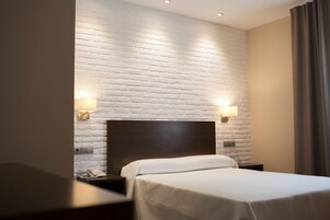 Habitación de hotel con pared de ladrillo blanco, cabecera de madera, cama con ropa de cama blanca, dos lámparas de noche y una mesita de noche con un teléfono.