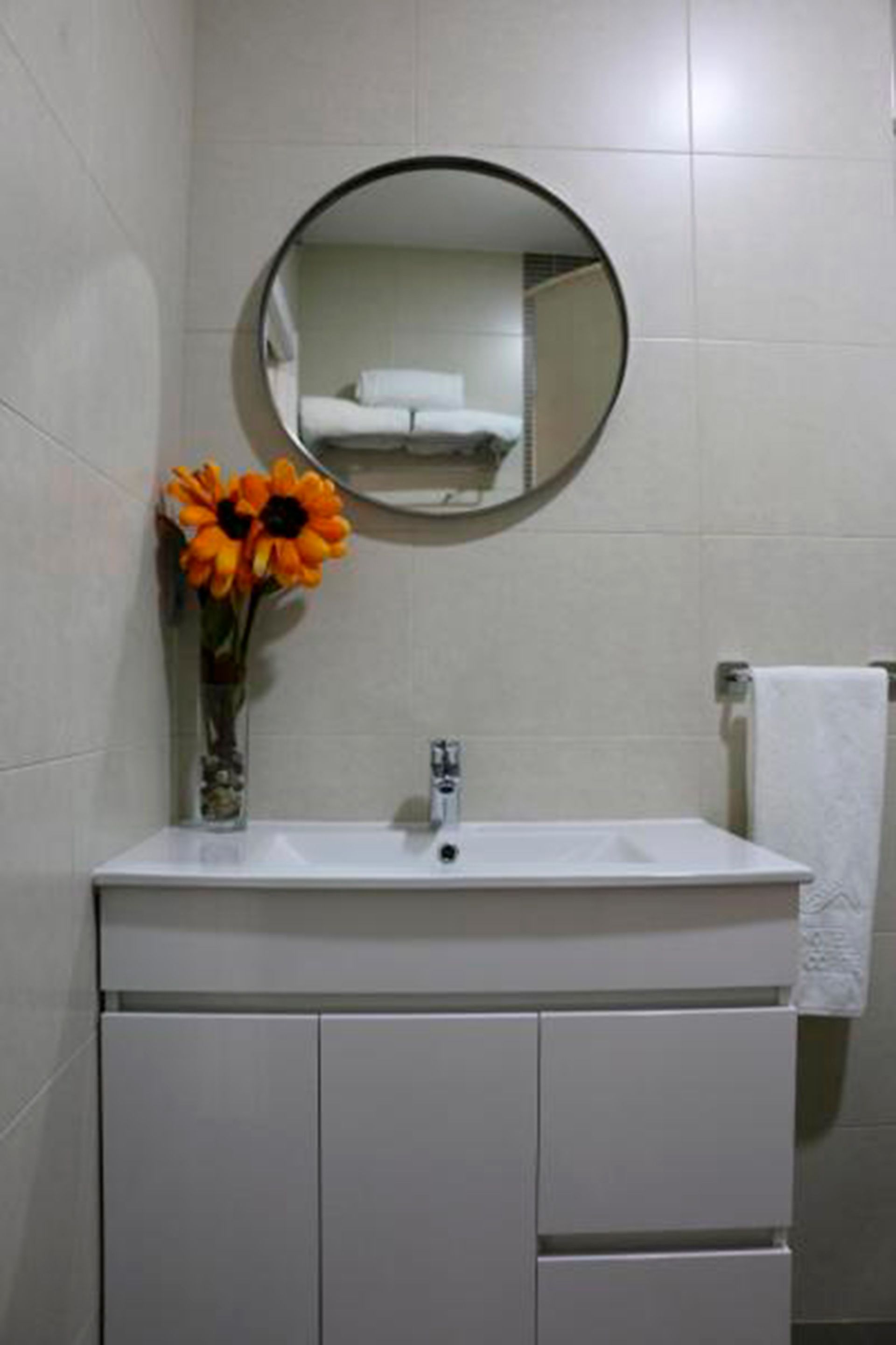 A bathroom with a white sink, a round mirror, and a vase with orange flowers.