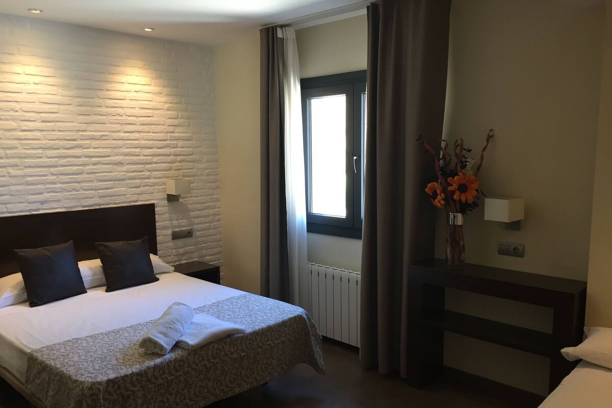 A hotel room with a bed, a nightstand, a window with curtains, a vase with flowers, and a wall-mounted light.