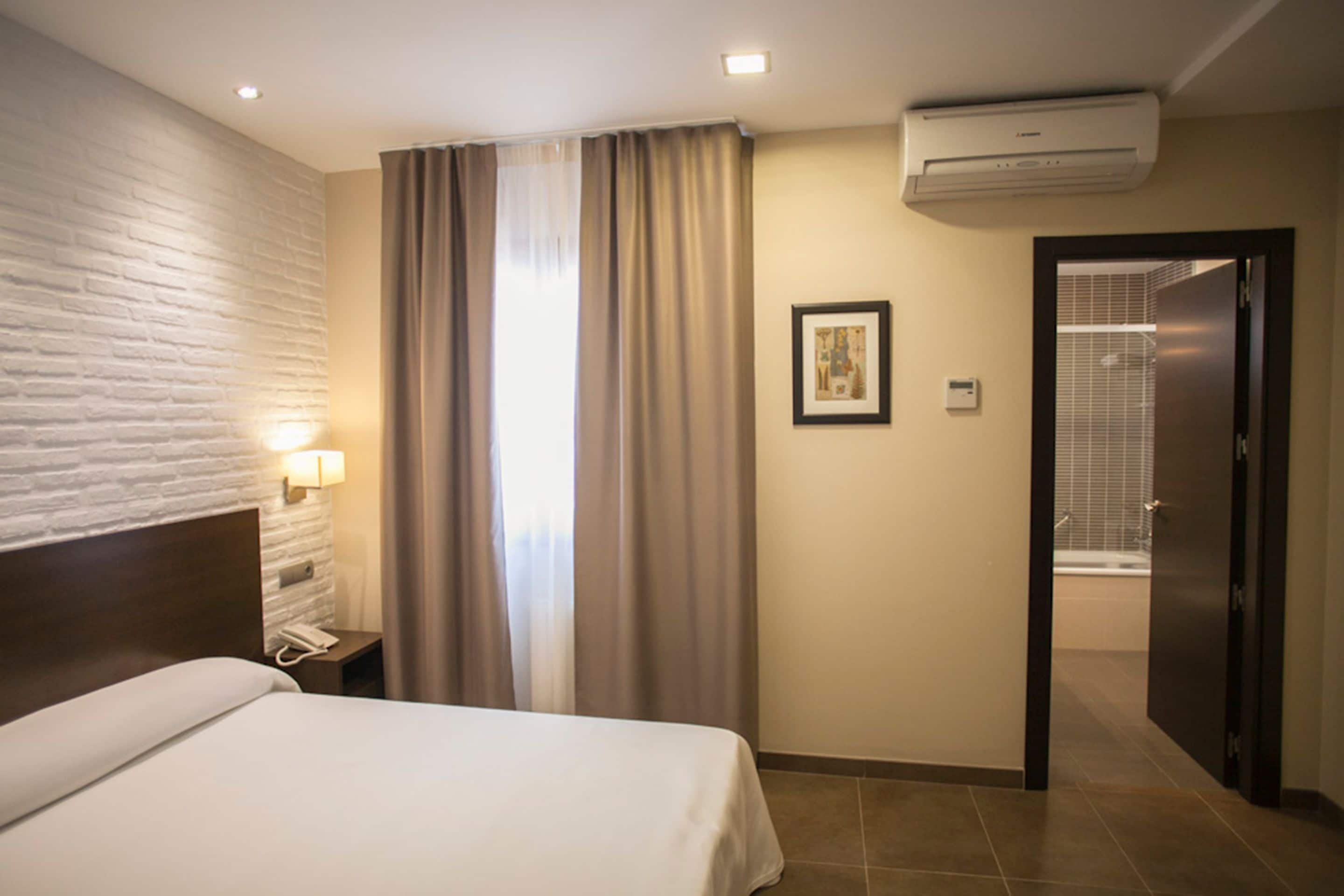 A hotel room with a bed, a nightstand, an air conditioning unit, a bathroom door, and a wall-mounted light fixture.