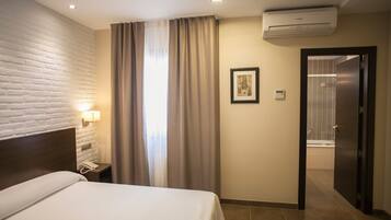 A hotel room with a bed, a nightstand, an air conditioning unit, a bathroom door, and a wall-mounted light fixture.