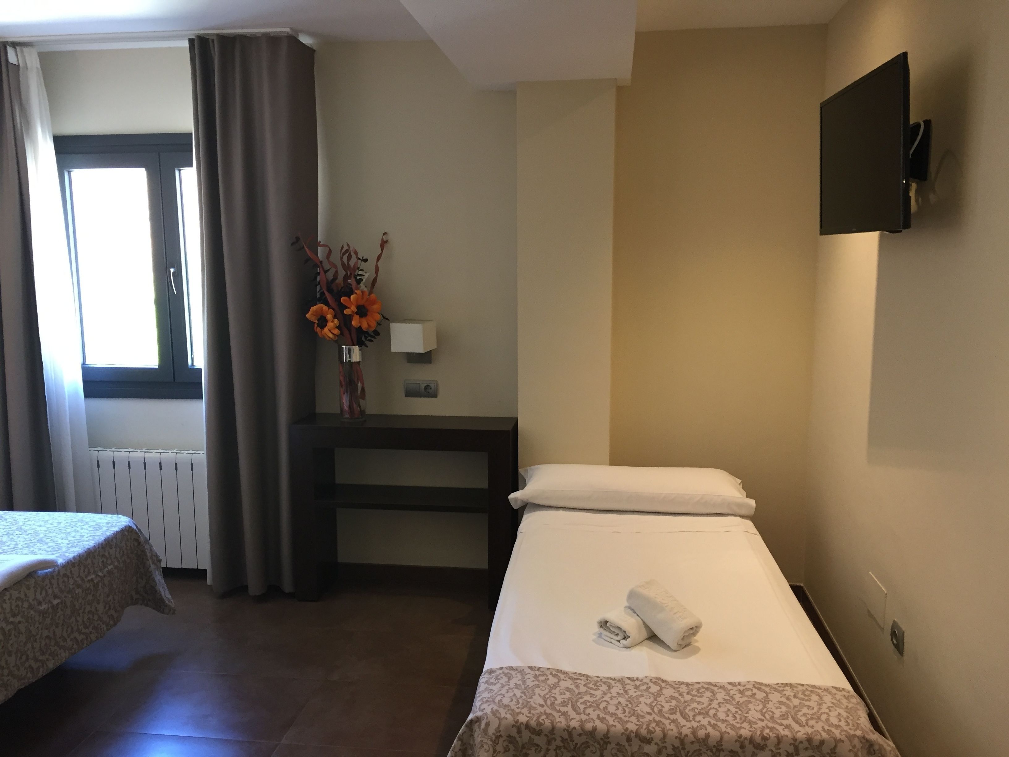 A hotel room with two beds, a desk with a vase of flowers, a TV mounted on the wall, and a window with curtains.