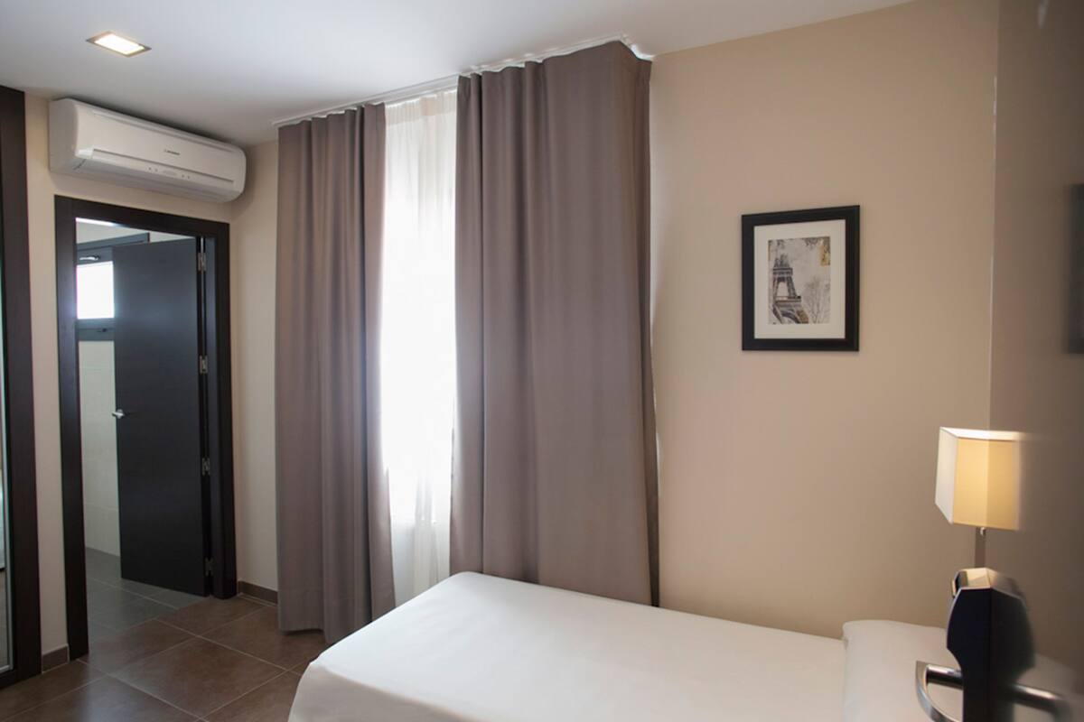 A hotel room with a bed, a desk, a lamp, an air conditioner, a door, and a window with curtains.