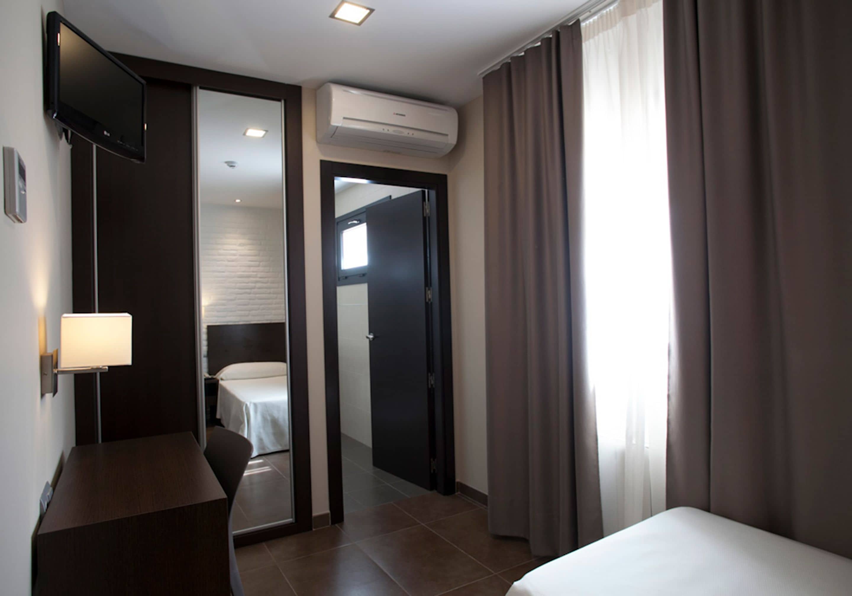 A hotel room with a bed, a desk, a TV mounted on the wall, and an air conditioning unit.