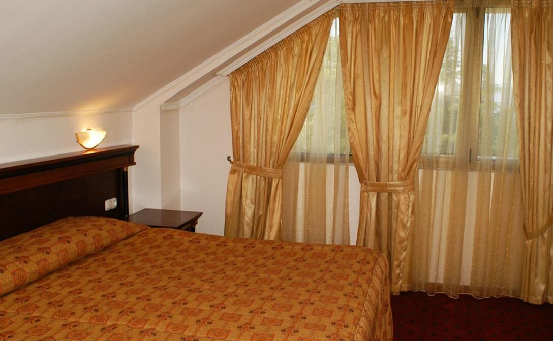 A room with a large bed, a nightstand, a wall-mounted lamp, and a window with curtains.