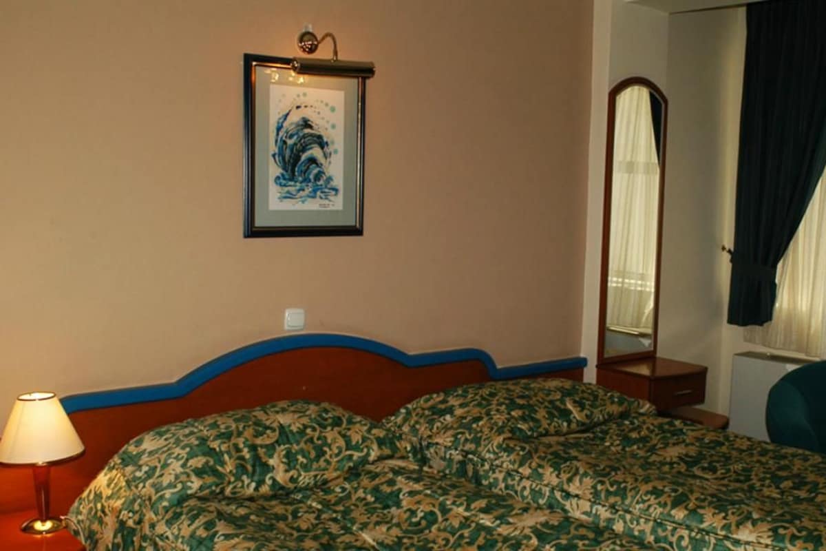 Quarto de hotel com cama, luminária, quadro na parede, espelho e janela com cortinas.