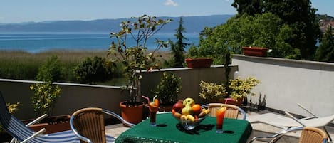 Una terraza con una mesa para dos, rodeada de sillas de mimbre, con vista al mar y las colinas de fondo.