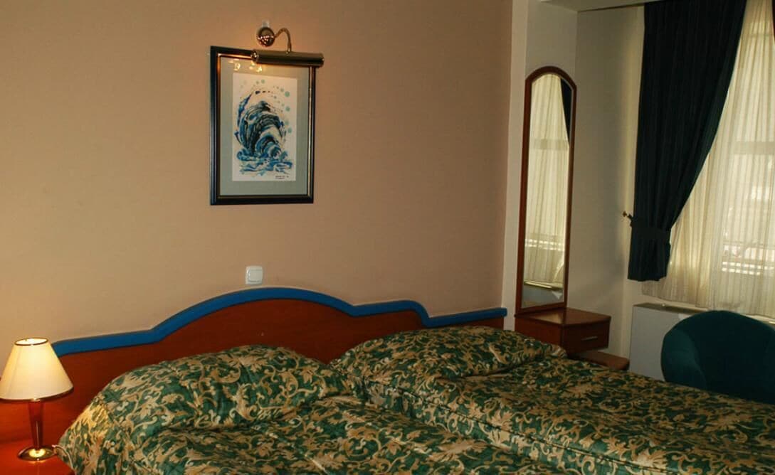 A hotel room with a bed, a bedside lamp, a framed artwork on the wall, a mirror, and a window with curtains.