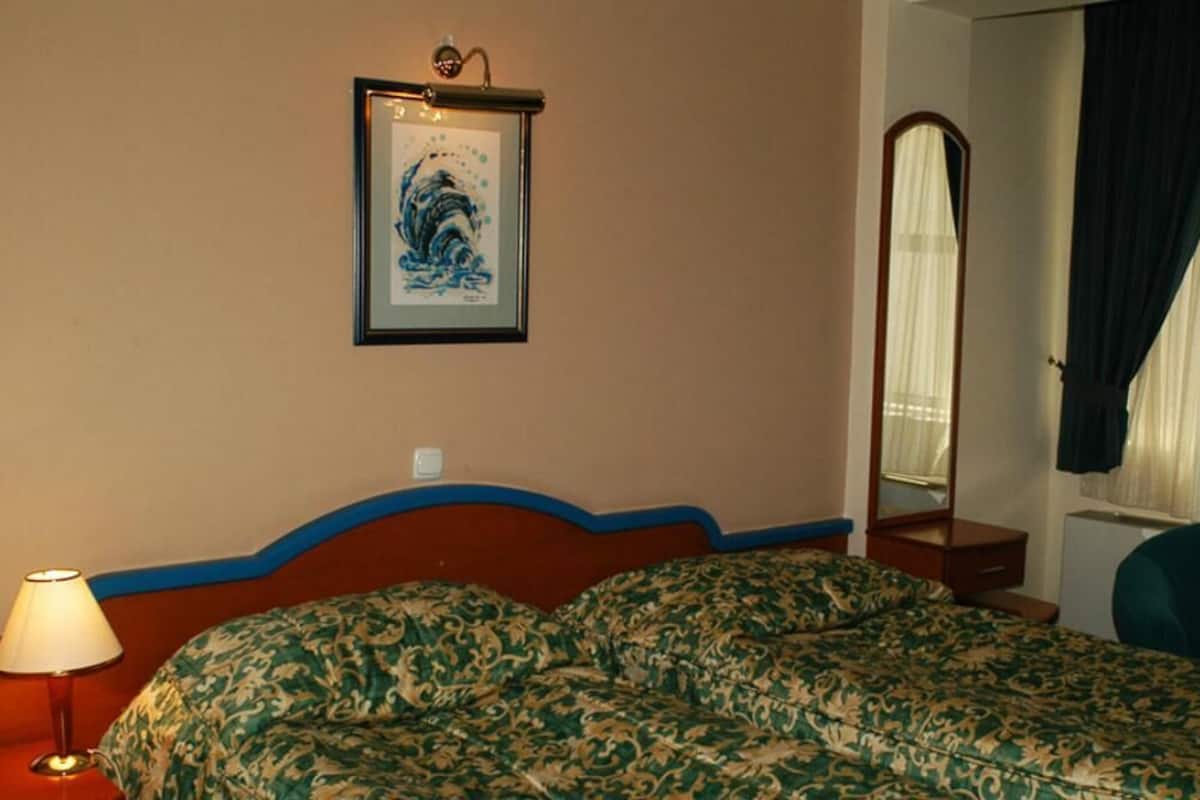 A hotel room with a bed, a bedside lamp, a framed artwork on the wall, a mirror, and a window with curtains.