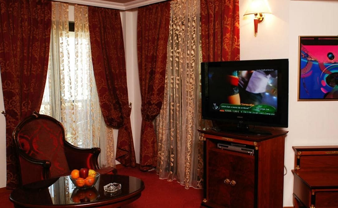 A room with a television on a wooden stand, a glass table with fruit, and curtains.