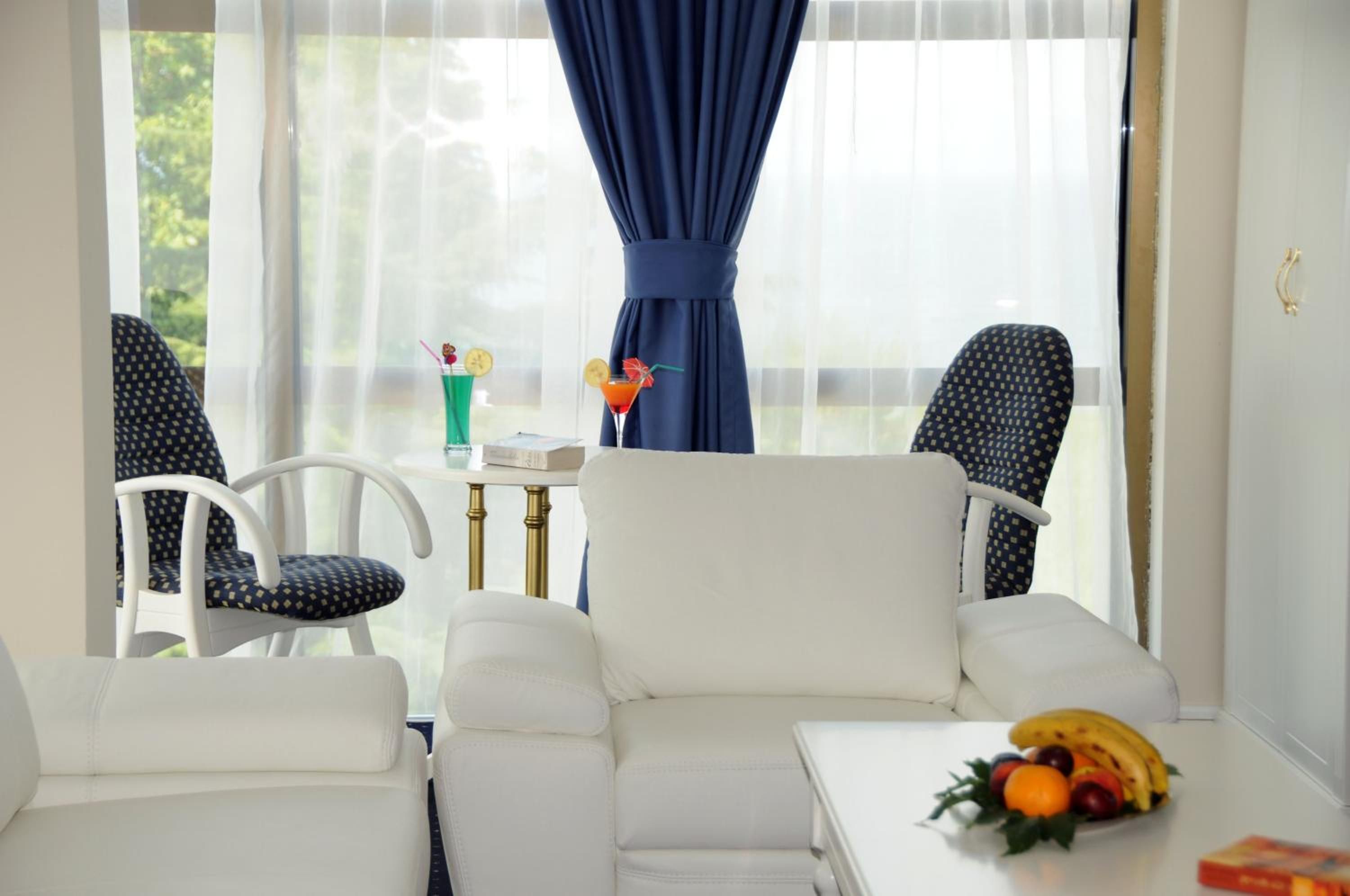 A room with a white sofa, two patterned chairs, a small table with a drink and fruit, and a view through sheer curtains.