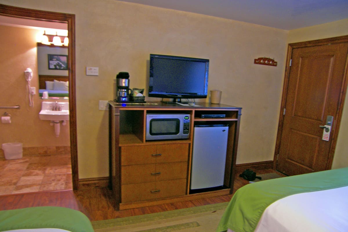 A hotel room with a wooden desk, a microwave, a small refrigerator, a television, a sink, and a toilet. There are two beds with green and white bedding.