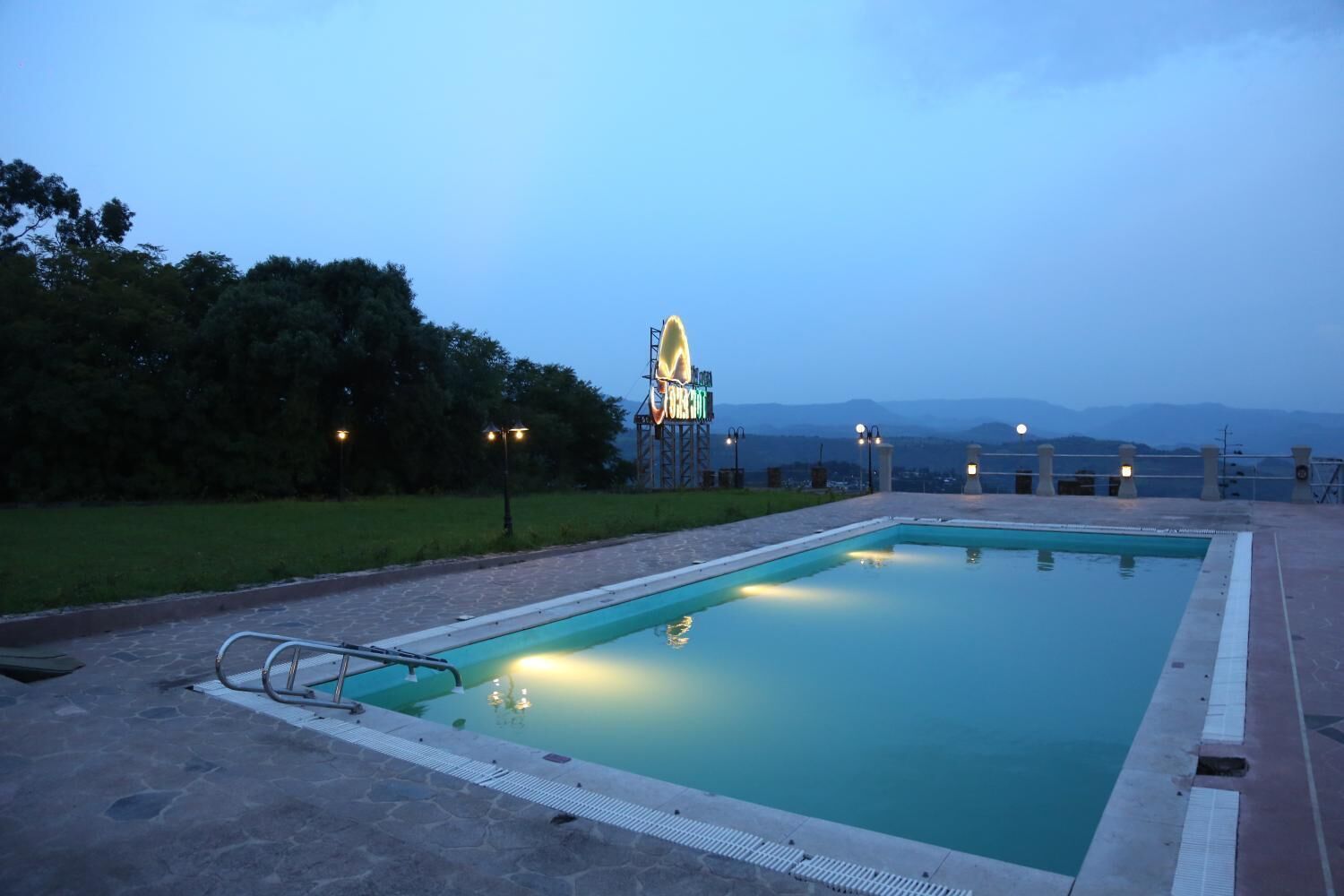 Una piscina rettangolare con una scala metallica su un lato, circondata da un solarium lastricato. Sullo sfondo, un grande cartello illuminato con la scritta “BAYVIEW” e vista di montagne in lontananza.