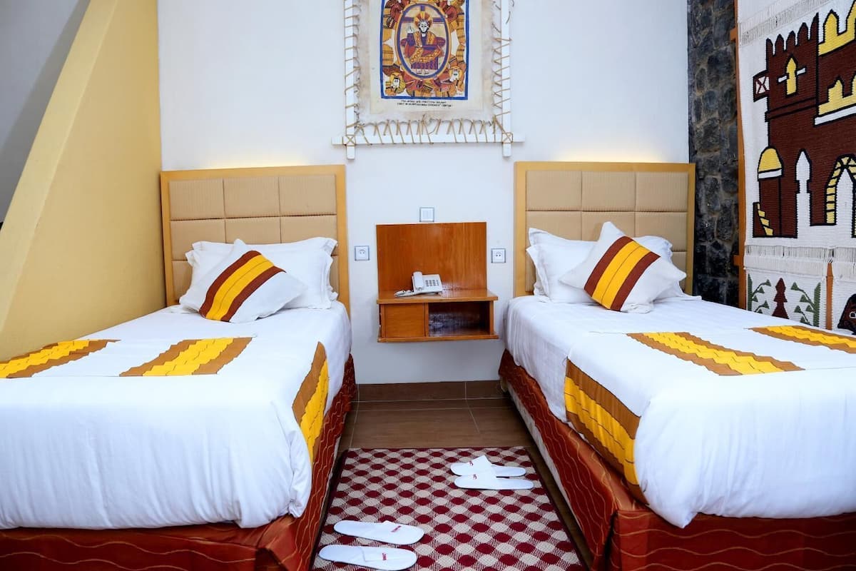 A hotel room with two beds, a checkered floor, a wooden nightstand, a telephone, and a decorative wall piece.