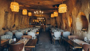 A restaurant with wooden tables and chairs, hanging chandeliers, and a stone wall interior. - Dere Suites (Ürgüp)