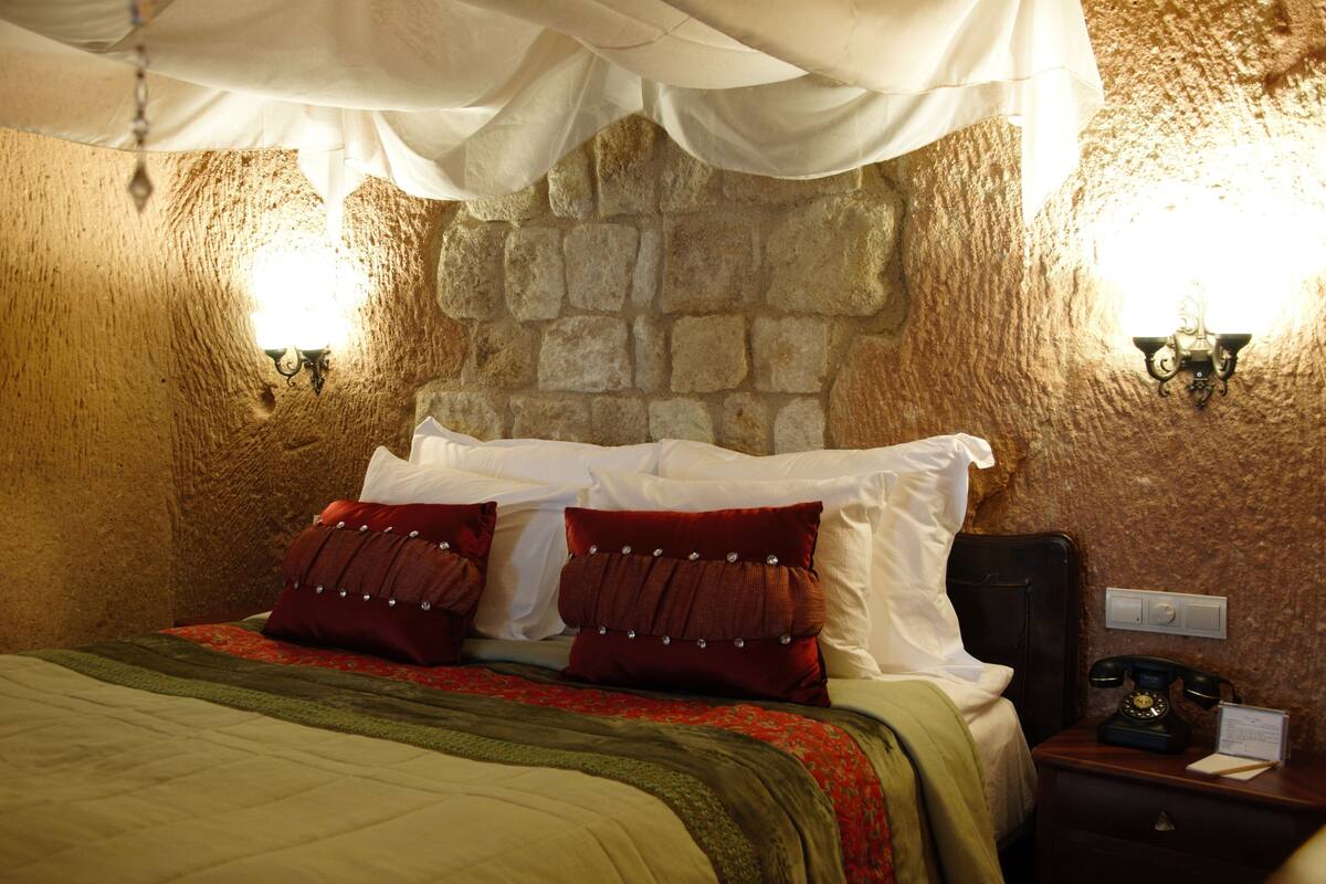 Una habitación con pared de piedra, una cama con almohadas blancas y rojas, una manta verde y una mesita de noche con teléfono.