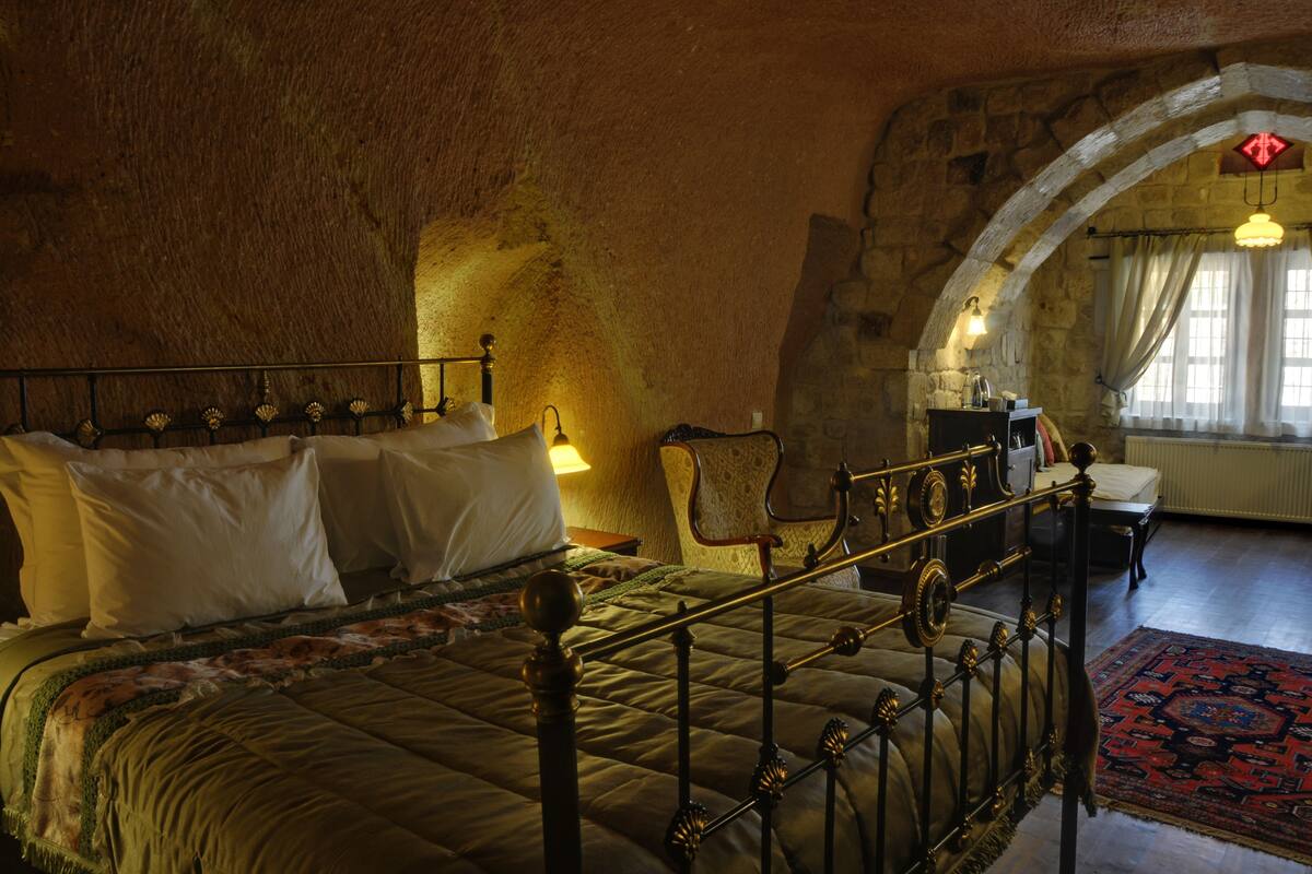 Una cama con estructura de metal, una silla y una mesa en una habitación con paredes de piedra y una ventana.
