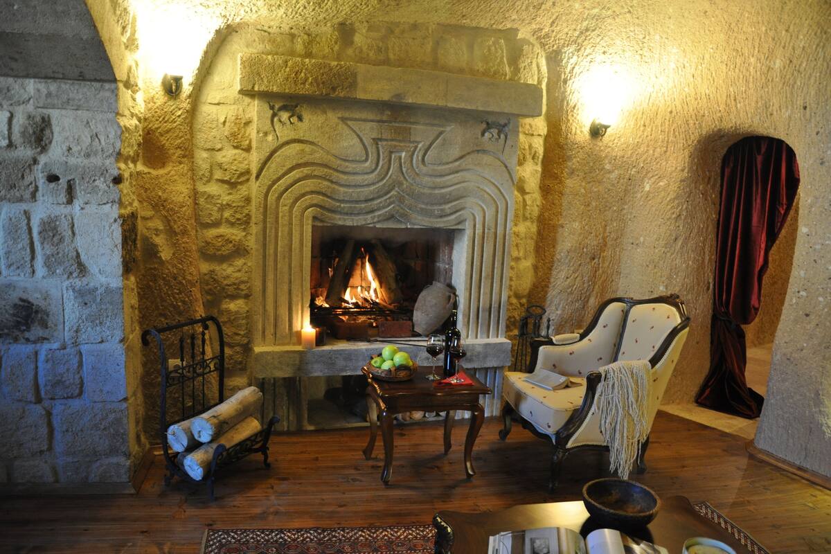 Una habitación acogedora con chimenea de piedra, una mesa de madera con fruta, una silla y un atizador de leña.