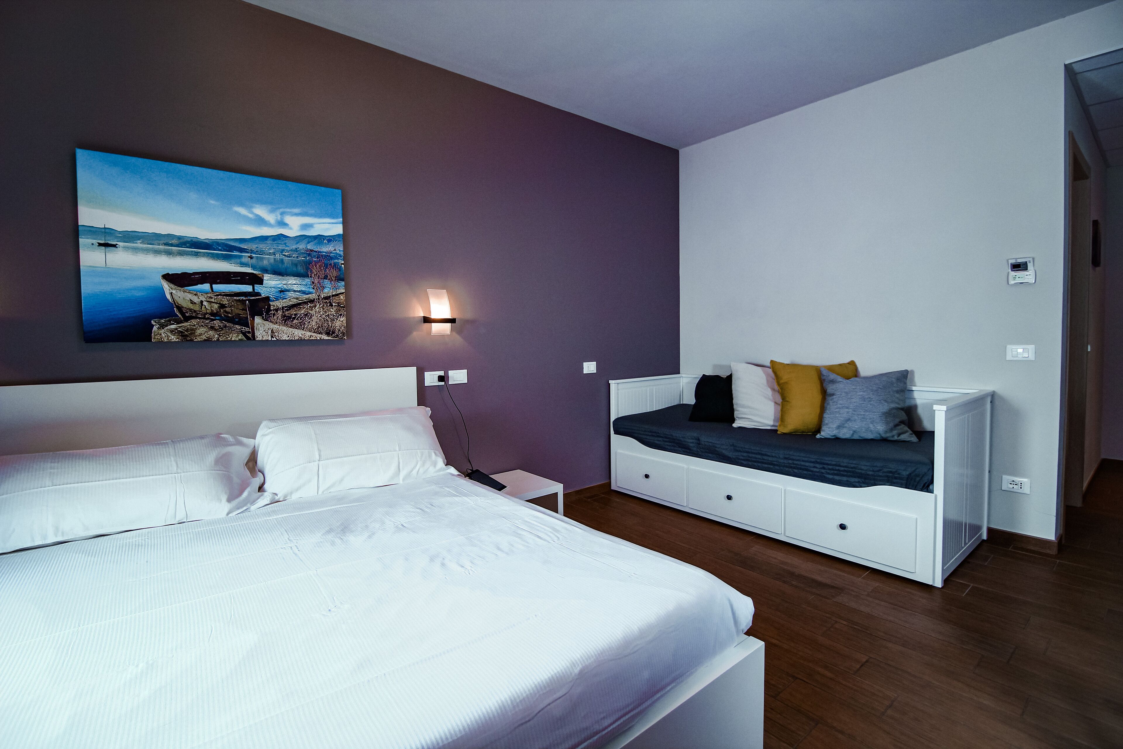 A bedroom with a large bed, a bedside table, a wall-mounted light, and a painting of a lake scene.