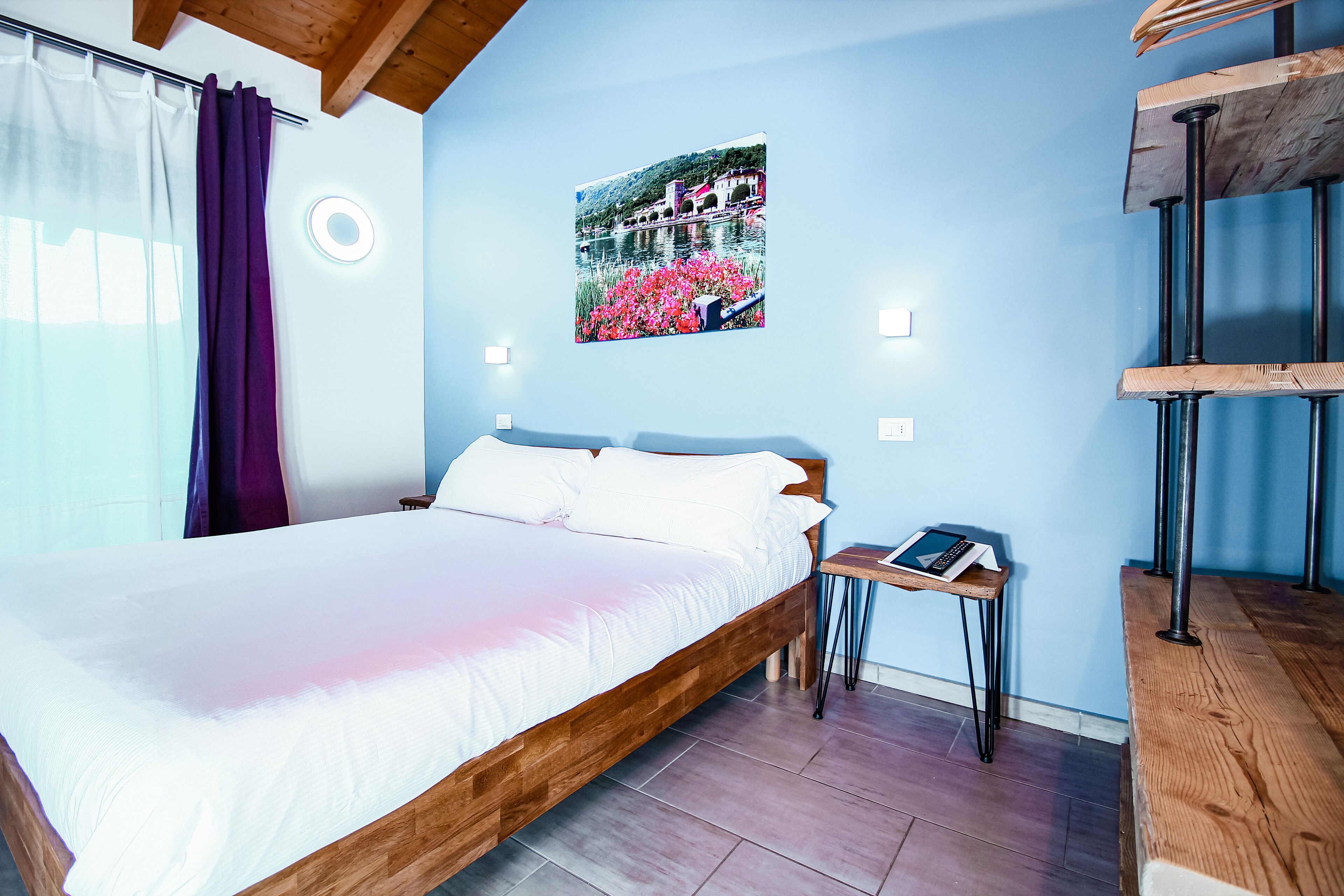 A bedroom with a wooden bed, a small bedside table with a book, a wall-mounted picture, and a wooden bench.