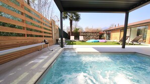 A swimming pool with a wooden deck and lounge chairs, surrounded by a garden and a modern building. - Agriturismo Il Cucchiaio di Legno (Orta San Giulio)