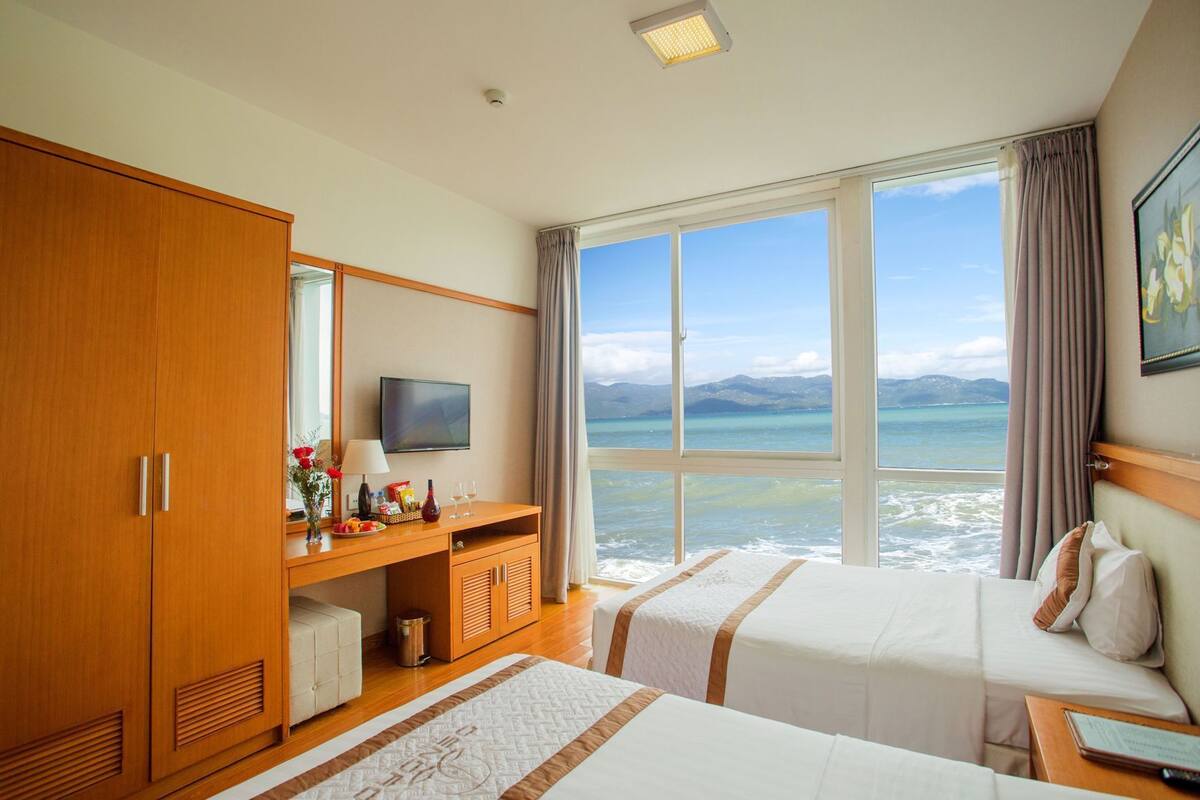 A hotel room with a bed, a desk with a TV, a wardrobe, and a large window with a view of the sea.