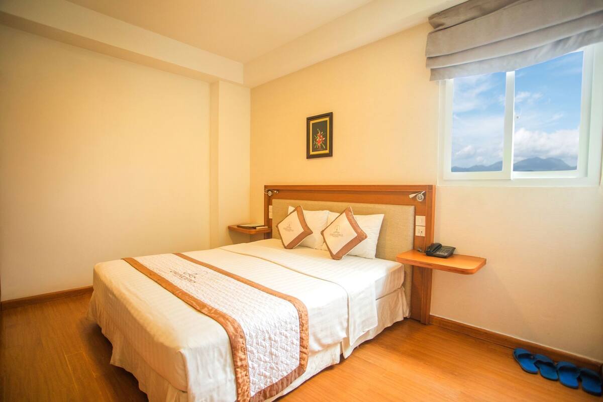 A neatly arranged bedroom with a bed, bedside tables, a nightstand, a window with a view, and a pair of slippers on the floor.