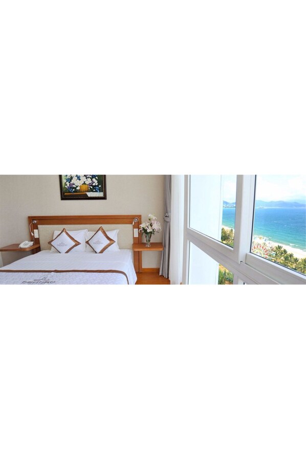 A hotel room with a large bed, a nightstand, a bedside table, a framed picture on the wall, and a window with a view of the beach and ocean. - Dendro Hotel (Nha Trang)