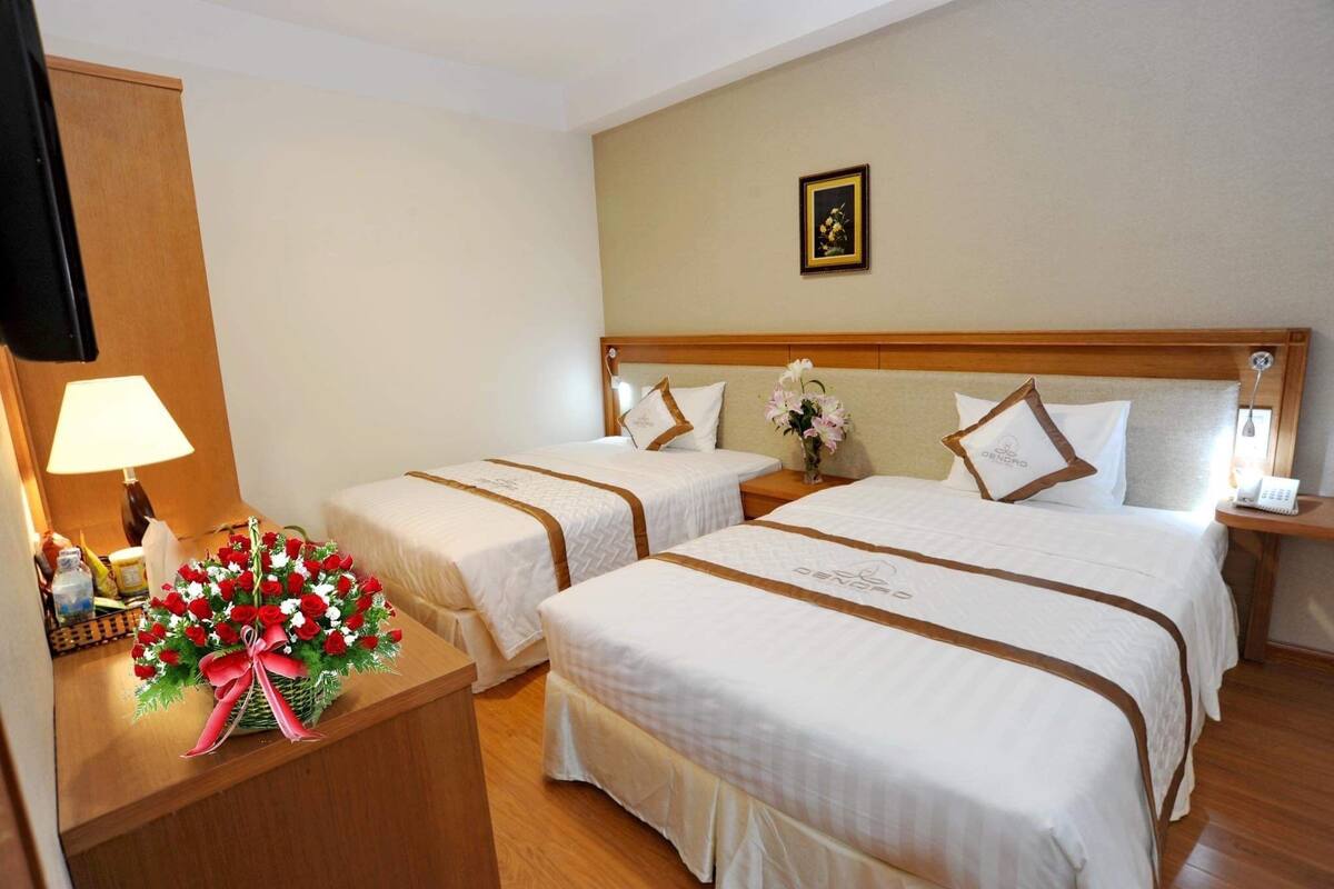 A hotel room with two beds, a wooden headboard, a bedside table with a lamp, a vase with flowers, and a framed picture on the wall.