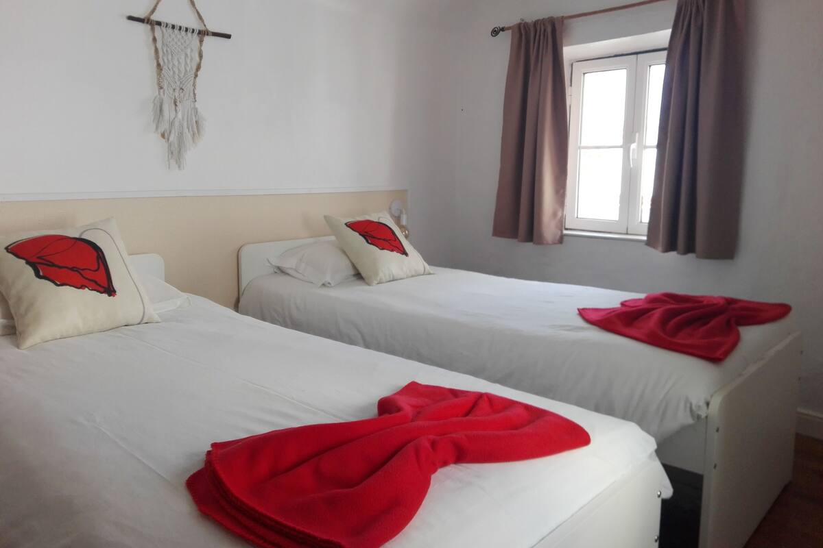 Dos camas individuales con ropa de cama blanca y almohadas rojas, un tapiz de macramé y una ventana con cortinas marrones.