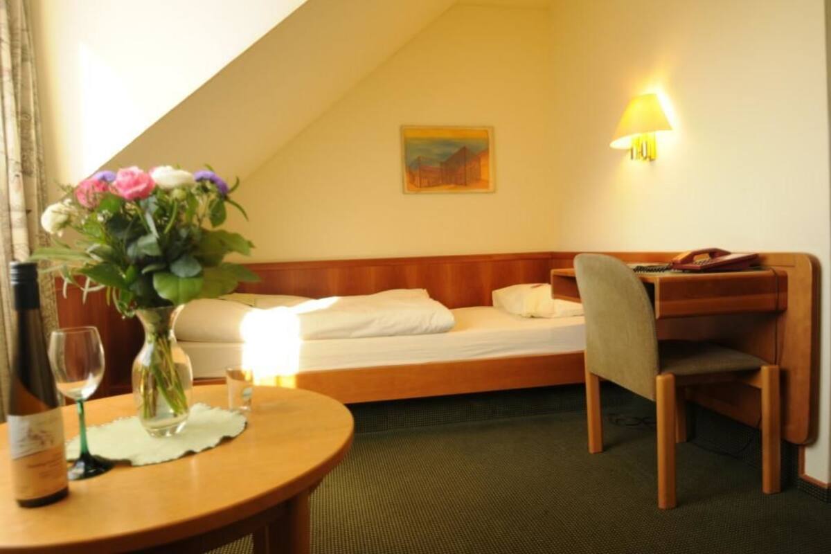 Et hotelværelse med seng, skrivebord, stol, lampe, vase med blomster, en vinflaske og et glas.