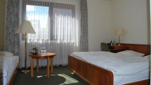 A hotel room with two beds, a round table with a lamp, and a view of the city through sheer curtains. - Hotel Rheingauer Tor (Hochheim am Main)