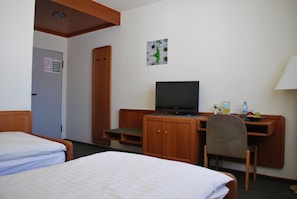 A hotel room with two beds, a desk with a computer, a chair, a TV, and a small table with a water bottle and fruit. - Hotel Rheingauer Tor (Hochheim am Main)