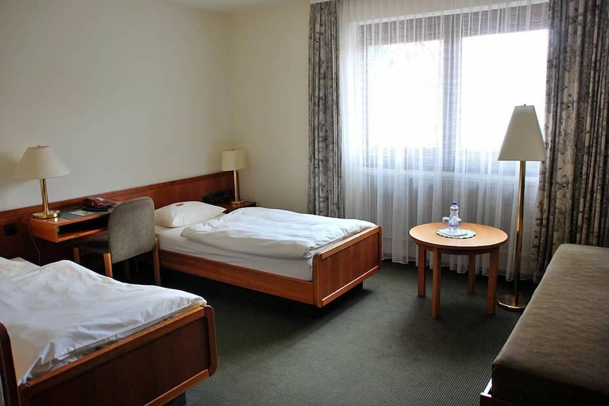 Et hotelværelse med to enkeltsenge, et skrivebord med lampe, et lille rundt bord og et stort vindue med let gardin.