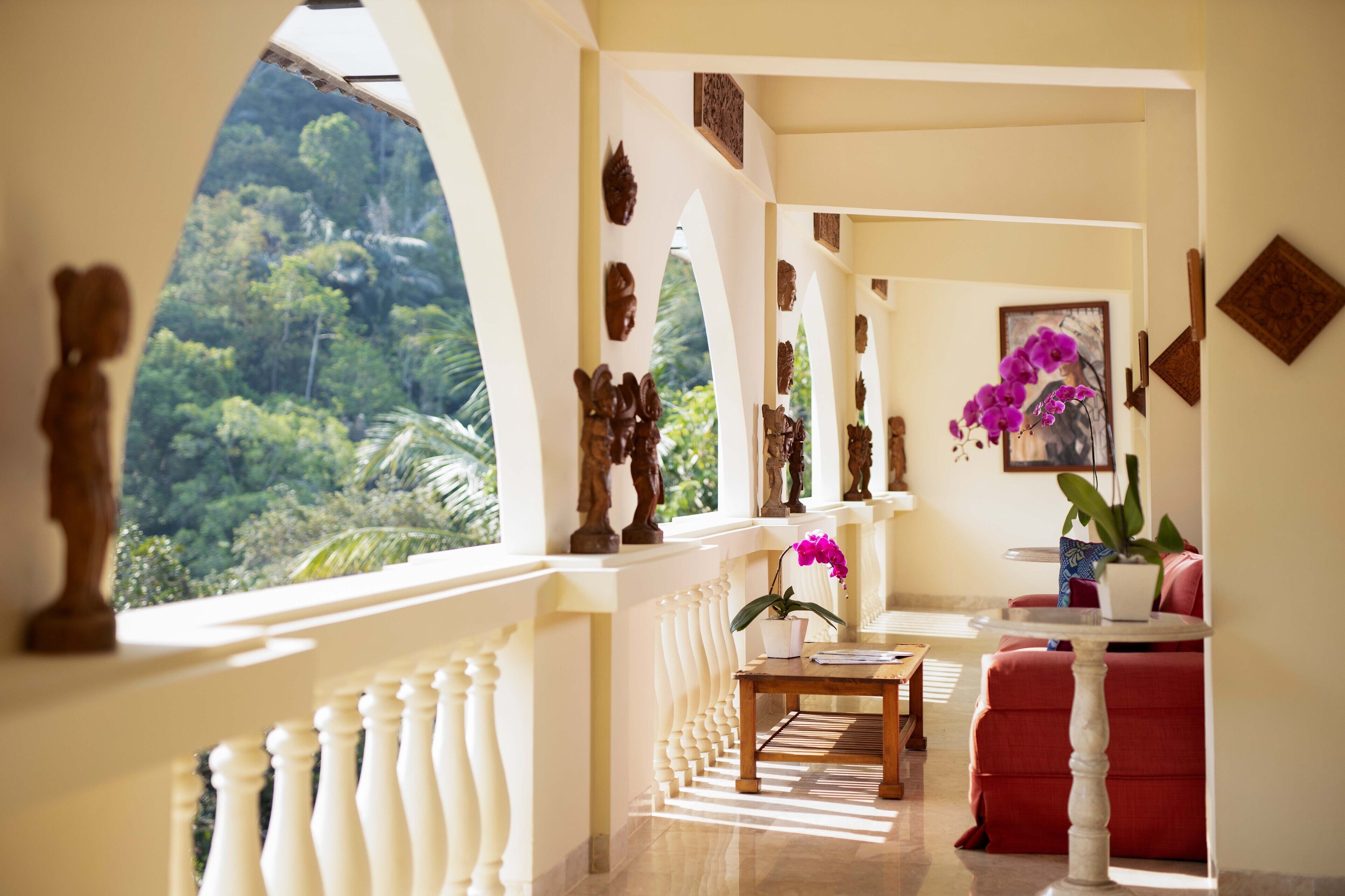 A balcony with a view of lush greenery, a wooden table with a vase of flowers, and a red sofa.