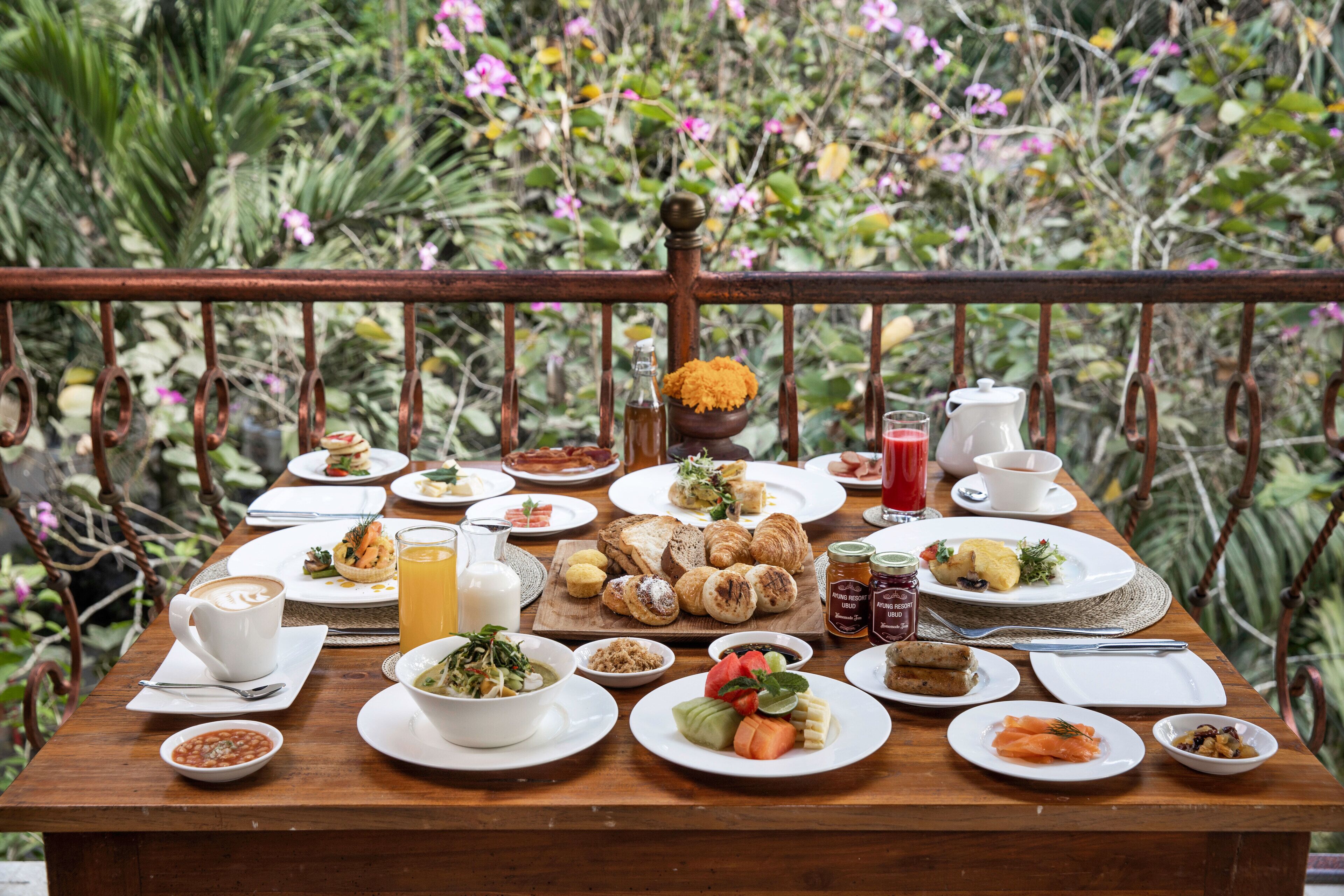 Una mesa de madera dispuesta con diferentes alimentos para el desayuno, como cruasanes, frutas y bebidas, en un balcón exterior con barandilla metálica y abundante vegetación de fondo.
