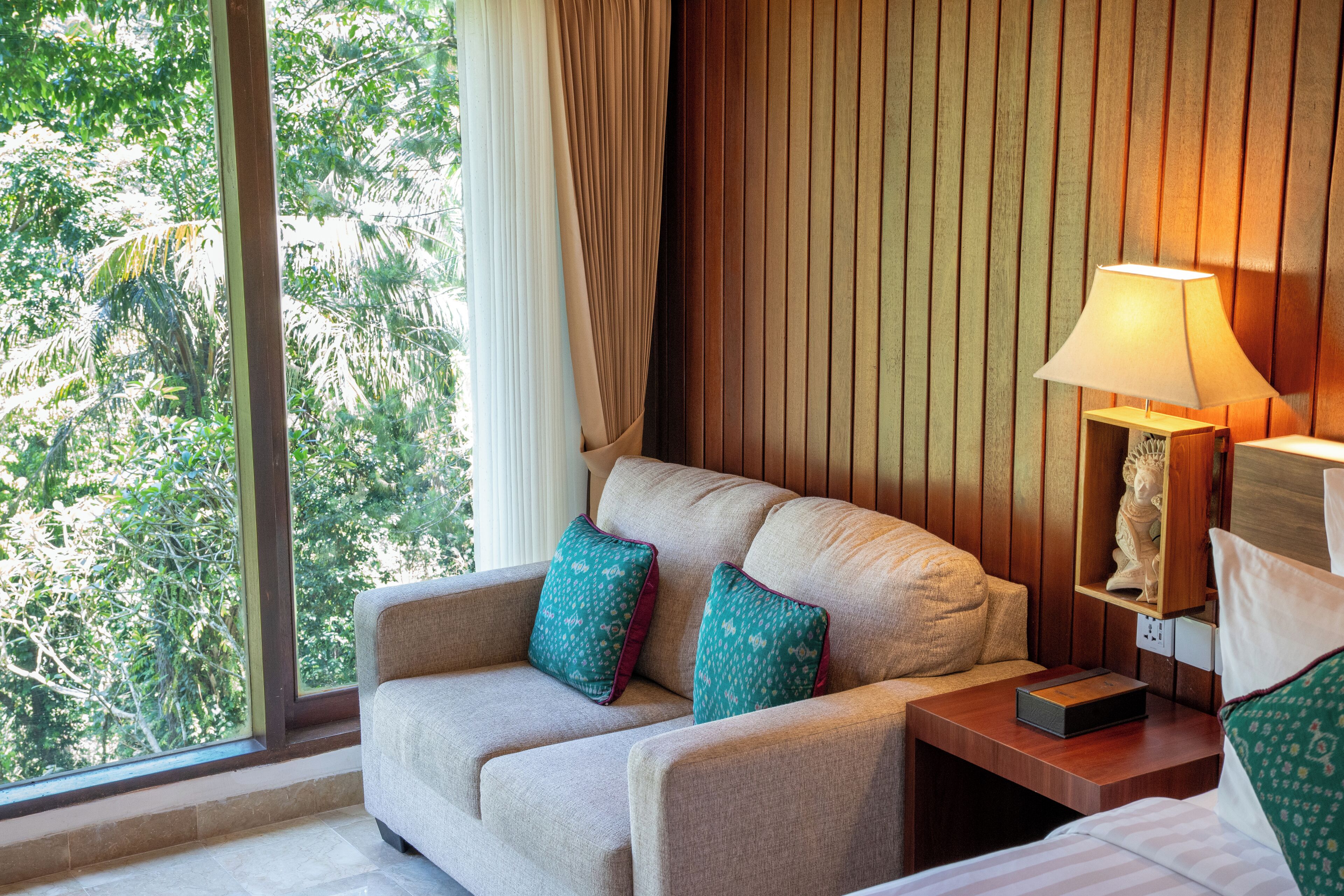 A hotel room with a sofa, a bedside table with a lamp, and a view of trees through a large window.