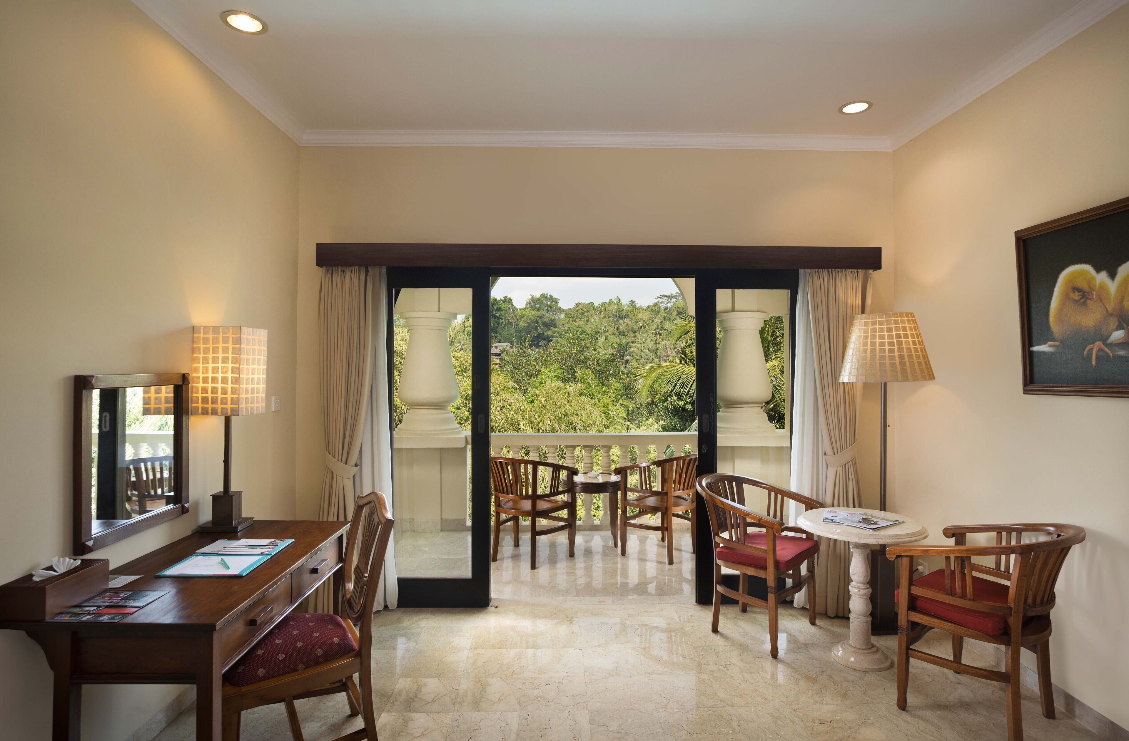 Habitación de hotel con balcón, muebles de madera, un escritorio con papeles y vista a zonas verdes.