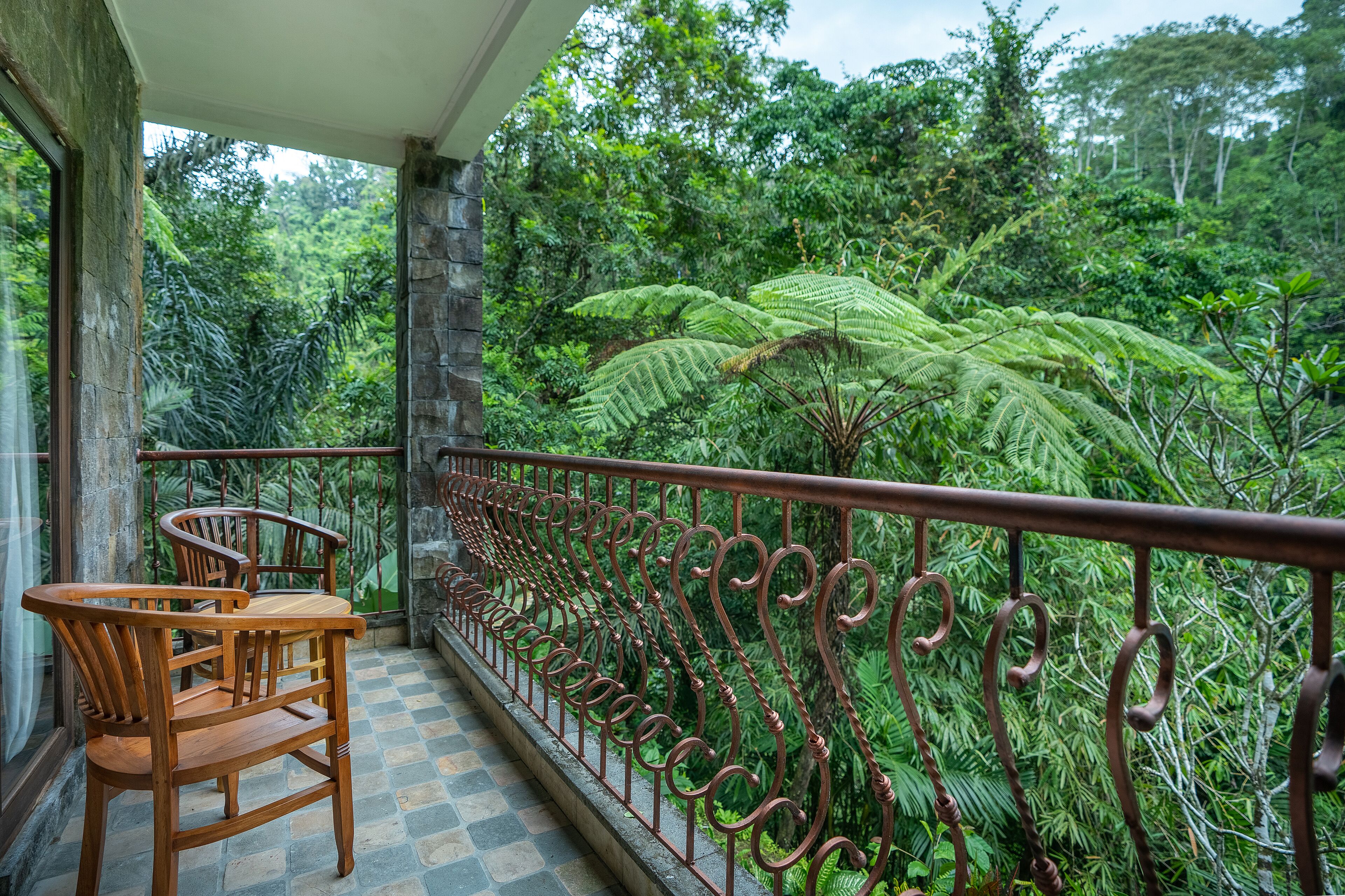 Un balcón con baranda de hierro forjado, sillas de madera y vistas a una exuberante vegetación tropical.