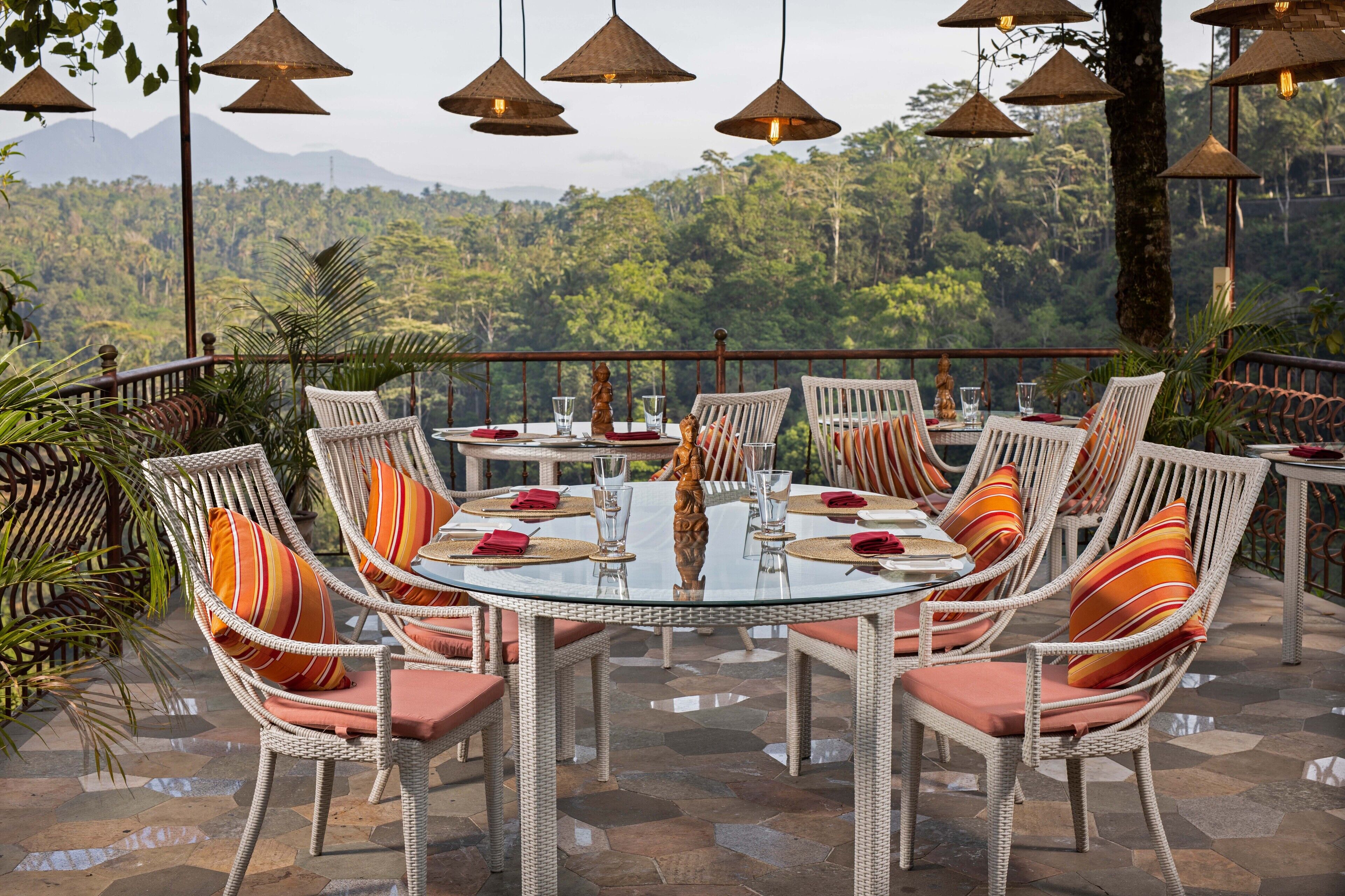 Un espace de restauration en plein air, avec des chaises en osier et une table en verre, entouré de plantes tropicales et offrant une vue panoramique sur les montagnes.