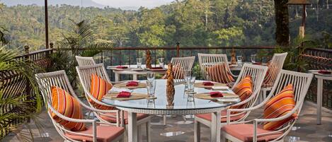 Un espace de restauration en plein air, avec des chaises en osier et une table en verre, entouré de plantes tropicales et offrant une vue panoramique sur les montagnes.