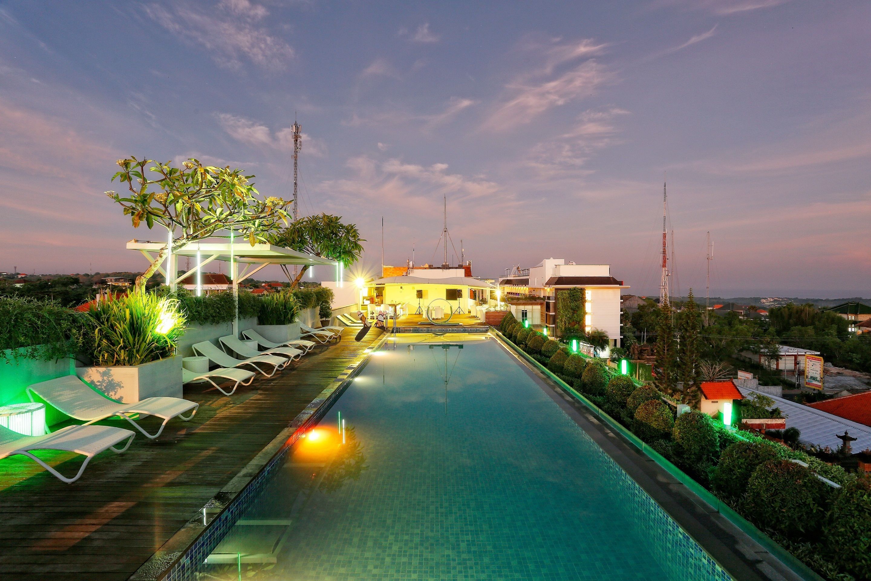 A rooftop swimming pool with lounge chairs and a pergola, surrounded by greenery and buildings.