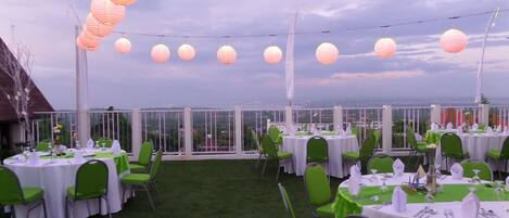 Un espacio de comedor al aire libre con mesas redondas, sillas verdes y luces colgantes.