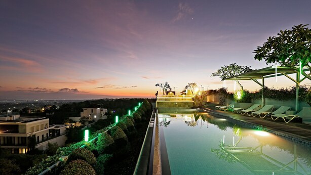 Una piscina infinita en la azotea con vistas a la ciudad al atardecer, rodeada de vegetación y sillas de descanso.