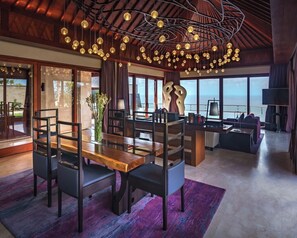 A spacious dining area with a wooden table and chairs, a large window with a view of the sea, and a ceiling with hanging spherical lights.