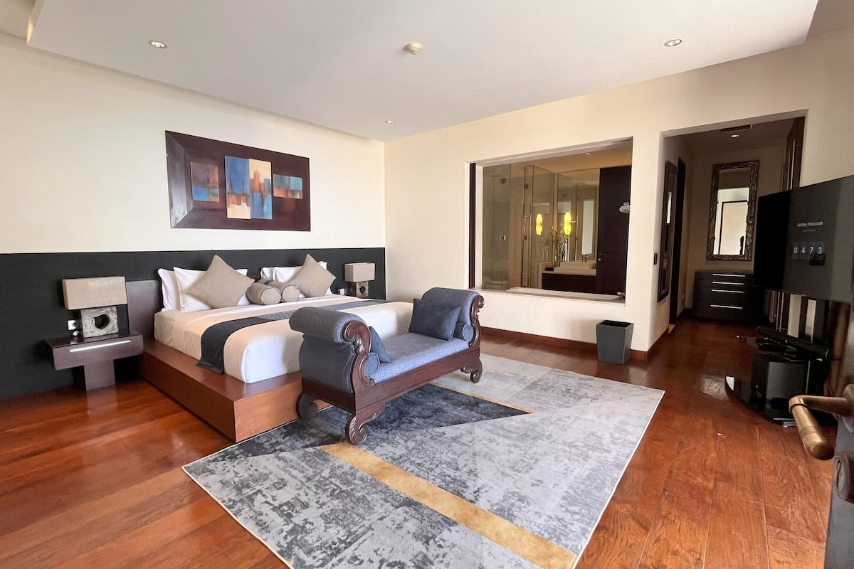 Una habitación de hotel moderna con una cama grande, un sofá, un televisor y un baño visible a través de una puerta abierta.