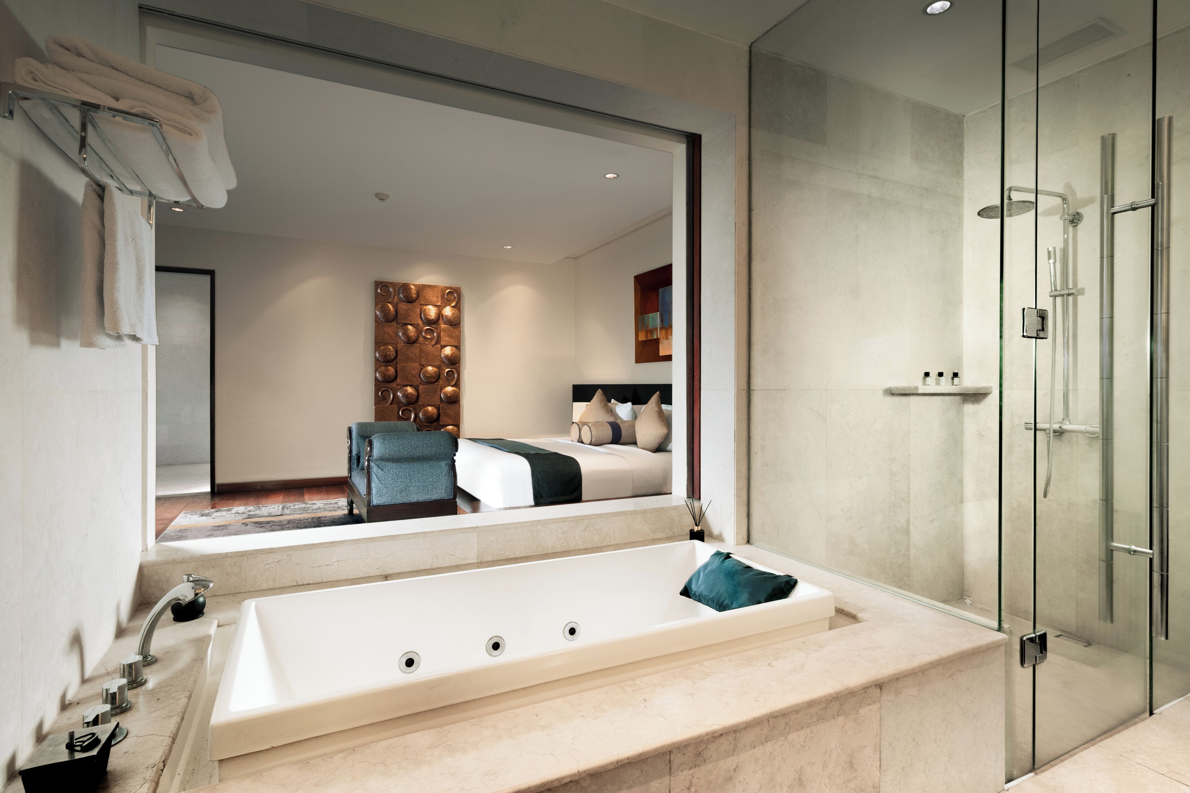 A modern bathroom with a large bathtub, a glass-enclosed shower, and a visible bedroom with a bed and a chair.