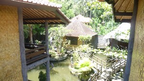 A traditional bamboo structure with a tiled roof, a small pond with a wooden bridge, and lush greenery in the background. - Kangkung Cottages (Karangasem)